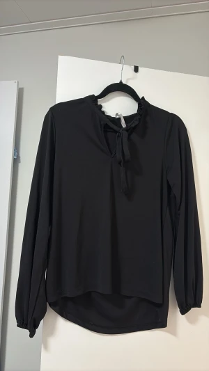 Svart blus med knyt och volang - Stilren svart blus med långa ärmar, volangdetalj vid halsringningen och knytband framtill. Blusen har en något lös passform och rundad nederkant. Perfekt att matcha med jeans eller kjol för en trendig look.