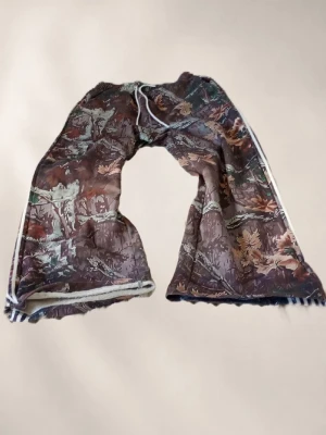 Camo mjukisbyxor med vita ränder - Säljer ett par mjukisbyxor med camomönster i brunt, grönt och beige. Byxorna har vita sidoremsor längs benen och dragsko i midjan. De är vida i modellen och har resår i midjan för extra komfort. Perfekta för en avslappnad och trendig look.