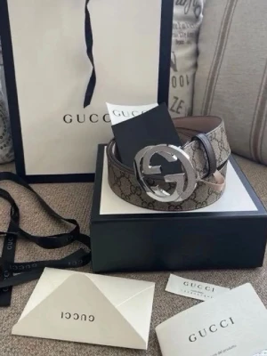 Gucci beige GG-bälte med silverdetalj - Säljer ett ikoniskt Gucci-bälte i beige med det klassiska GG-mönstret och en stor, rund silverfärgad GG-spänne. Bältet har bruna detaljer och är tillverkat i skinn och syntet. Perfekt accessoar för att lyfta vilken outfit som helst.