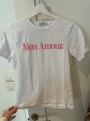 Vit t-shirt Mon Amour från Stradivarius - Vit t-shirt från Stradivarius med trycket 'Mon Amour.' i rosa framtill. Klassisk rund halsringning och korta ärmar. Perfekt basic.