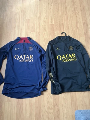 PSG Bundel - Två snygga PSG träningsjackor, en marinblå från Nike med röd insida på kragen och en svart från Jordan med gula detaljer. Båda har Qatar Airways-tryck, halv dragkedja och klubbmärke på bröstet. Perfekta för fotbollsträning eller chill.