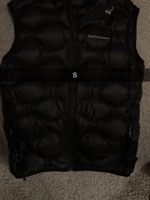 Svart dunväst Peak Performance S - Snygg svart dunväst från Peak Performance med dragkedja framtill och en bröstficka med dragkedja. Västen har diskret logga på bröstet och quiltad design som ger en sportig look. Perfekt att slänga över en hoodie eller tröja för extra värme. Ny pris 1199, säljer för 499 ny skick