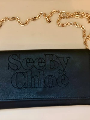 See by Chloé wallet on chain – som ny ✨  - See by Chloé – svart långplånbok i pebbled läder med guldkedja  Mycket chic See by Chloé continental wallet i svart pebbled läder. Kan bäras som plånbok, clutch eller crossbody-väska med den medföljande guldkedjan.  Märke: See by Chloé (diffusionslinje från det franska modehuset Chloé) Modell: Långplånbok / continental wallet Material: Pebbled läder Färg: Svart Mått: 19 × 10,5 cm Stängning: Guldtonat blixtlås med märkt dragsko Interiör: Sedelfack, kreditkortsfickor, textil Exteriör: Äkta läder