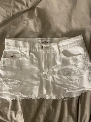 Vit jeanskjol från Hollister - Vit kort jeanskjol från Hollister med slitna detaljer och rå kant nertill. Klassisk femficksmodell med bälteshällor och dragkedja fram. Märke på 3dje bilden från tidigare ägare, jag har inte testat få bort det 