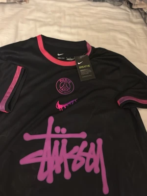 Svart PSG x Stüssy tröja Nike - Säljer en svart PSG x Stüssy tröja från Nike med rosa och lila detaljer. Tröjan har PSG-logga och Stüssy-grafitti framtill samt rosa Nike swoosh. Tillverkad i lätt och ventilerande material, perfekt för sport eller streetwear.
