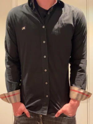 Svart skjorta från Burberry - Stilren svart skjorta från Burberry med klassiska rutiga detaljer på insidan av manschetterna och liten logga på bröstet. Skjortan har lång ärm, knappar framtill och en normal passform. Perfekt för dig som gillar en clean look med exklusiv touch.
