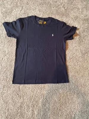 Ralph Lauren T-shirt – Mörkblå med Vit Logga (Storlek M) - Säljer en helt ny och oanvänd Ralph Lauren T-shirt i storlek M. Stilren och tidlös T-shirt i mörkblå färg med den klassiska vita Ralph Lauren-loggan broderad på bröstet. Tillverkad i mjukt och bekvämt material av hög kvalitet – perfekt för både vardag och en avslappnad, stilren look. 🔹 Märke: Ralph Lauren 🔹 Modell: T-shirt 🔹 Färg: Mörkblå med vit logga 🔹 Storlek: M 🔹 Skick: Helt ny / oanvänd 🔹 Passform: Klassisk Bara att höra av dig vid frågor eller funderingar 