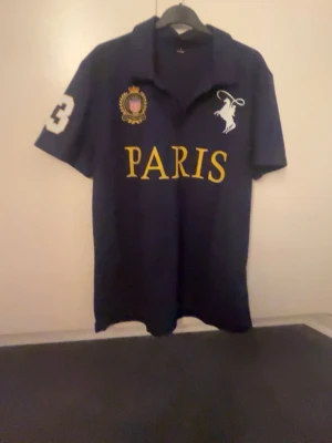 Marinblå pikétröja Paris Polo - Snygg marinblå pikétröja från Polo Ralph Lauren med stort gult PARIS-tryck på bröstet, vit polospelare och broderad patch. Nummer 3 på ärmen. Klassisk krage och knappar framtill. Perfekt för dig som gillar streetway stil.