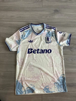 Aston Villa Adidas matchtröja - Säljer en snygg Aston Villa matchtröja från Adidas i vitt med ljusblå och ljusrosa detaljer. Tröjan har klubbmärke, Adidas-logga och Betano-tryck på bröstet samt blå ränder på axlarna. Tillverkad i ett lätt och ventilerande material, perfekt för fotboll eller träning.