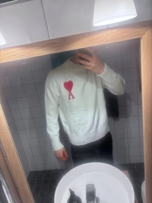 Vit stickad tröja från Ami Paris L - Säljer en vit stickad tröja från Ami Paris med röd hjärtlogga. Tröjan har rund halsringning, långa ärmar och ribbade muddar vid ärmslut och nederkant. Använt ett fåtal gånger.