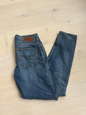 Blå jeans - Säljer ett par klassiska blå jeans med rak passform och lätt tvättad look. Jeansen har fem fickor, normal midja och bruna detaljer vid linningen. Perfekta för en avslappnad och stilren outfit.