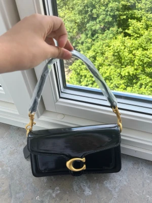 Svart lackad handväska från Coach - Snygg svart handväska i lackat läder från Coach med guldfärgade metalldetaljer och ett stort C-spänne framtill. Kort axelrem med plastskydd kvar. Perfekt till dig som vill ha en trendig och stilren accessoar. Långt band ingår