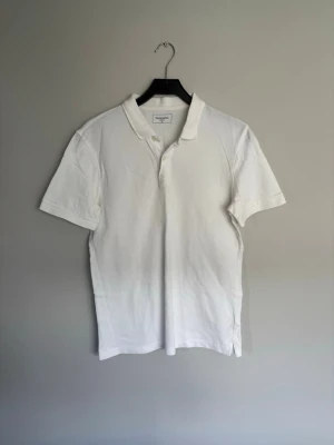 Vit pikétröja från Dressmann - Vit pikétröja från Dressmann i klassisk modell med krage och knappar framtill. Tröjan är tillverkad i bomull och har korta ärmar. Perfekt för dig som gillar en clean och enkel stil.
