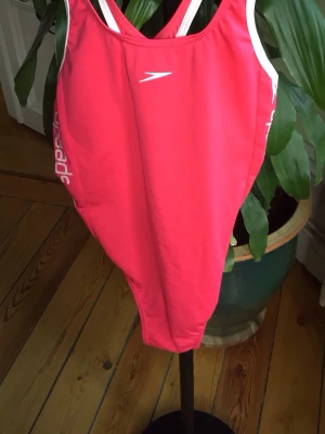 Röd Speedo baddräkt med logga - Röd baddräkt från Speedo med vita logotyper längs sidorna och öppen rygg. Tillverkad i Endurance-material som är känt för att vara extra tåligt och klorresistent. Perfekt för simträning eller strandliv.
