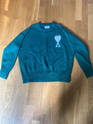 Grön stickad tröja från Ami Paris - Snygg grön stickad tröja från Ami Paris med deras ikoniska vita hjärta och A-logga på bröstet. Tröjan har rund hals, långa ärmar och breda ribbade muddar vid ärmslut och nederkant. Perfekt för en trendig och avslappnad look.