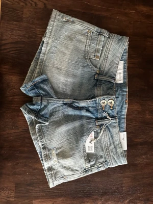 Ljusblå denimshorts från Zara - Säljer ett par ljusblå denimshorts från Zara med klassisk femficksdesign, råa kanter och dubbla knappar i midjan. Shortsen har en avslappnad passform och är perfekta för varma dagar. Ny pris  300