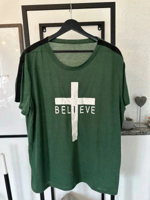 Grön t-shirt med tryck 'BELIEVE' - Snygge grön t-shirt med vit text och kors-tryck 'BELIEVE' på bröstet. T-shirten har svarta detaljer längs axlarna och en klassisk rund halsringning. Perfekt för dig som gillar statement-plagg och enkel stil.