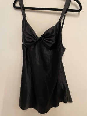 Svart satin klänning med rosett - Snygg svart kort klänning i satin med smala axelband och en stor rosett framtill vid bysten. Klänningen har en lätt glansig finish och en feminin siluett som ger en elegant look. Avklipp så en jämna kanter men rak 