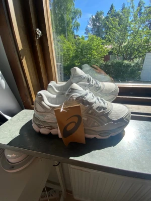 Grå Asics sneakers med mesh och mocka - Snygga grå sneakers från Asics med klassisk siluett. Skorna har ovandel i mesh och mocka för en sportig vibe och bra andningsförmåga. Tjock, dämpad sula i vitt och grått ger skön komfort. Perfekta för dig som gillar stilrena och trendiga sneakers.