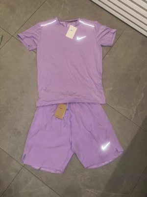 Lila träningsset från Nike - Snyggt lila träningsset från Nike med t-shirt och shorts. Båda plaggen har reflekterande Nike-logga och är gjorda i ett lätt, snabbtorkande material som passar perfekt för träning. T-shirten har rund hals och shortsen har dragsko i midjan.