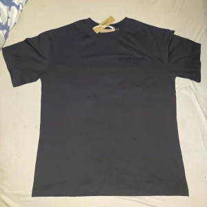 Svart Essentials Fear of God t-shirt - Säljer en svart t-shirt från Fear of God Essentials med stor logga på ryggen och diskret tryck fram. Klassisk passform och rund halsringning. Perfekt för dig som gillar streetwear och enkel stil.