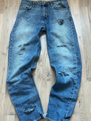 Vintage archive Blå slitna jeans med hål - Säljer ett par blå jeans med flera slitningar och hål för en riktigt avslappnad och trendig look. Jeansen har rak passform, klassisk femficksdesign och råa kanter vid bensluten. Perfekta för dig som gillar en edgy streetstil.