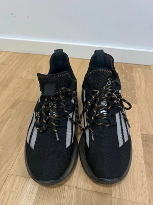 Svarta emporia Armani  - Snygga svarta sneakers med vita ränder på sidorna och orange detaljer. Skorna har stickad ovandel, tjock sula och svarta snören med orange inslag. Perfekta för dig som gillar sportig stil och vill ha något bekvämt till vardags.