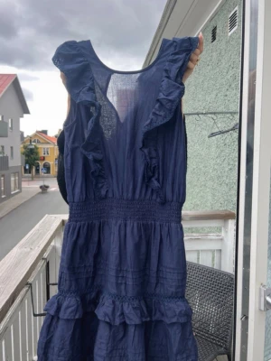 Marinblå volangklänning i bomull - Hej jag säljer en blå klänning med detaljer och volanger vid axlarna och nedåt kjoldelen. Klänningen är i ett väldigt bra skick inga fläckar då den är använd en gång tidigare.