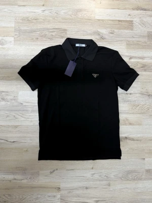 Svart pikétopp från Prada - Klassisk svart pikétopp från Prada med krage och korta ärmar. Tidlös design med diskret logga på bröstet och knappslå vid halsen. Perfekt för en stilren och avslappnad look.