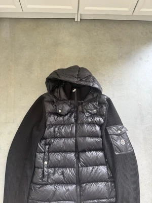 Moncler cardigan - Snygg svart dunväst från Moncler med stickade ärmar och huva. Västen har dragkedja framtill, fickor med tryckknappar och en ikonisk Moncler-logga på ärmen. Materialet är en mix av dun och stickat tyg för extra komfort och stil.