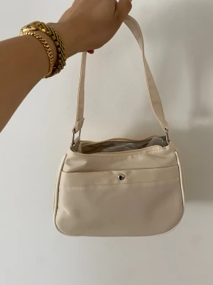 Beige axelväska från Wivdora - Snygg beige axelväska från Wivdora med enkel, stilren design. Väskan har en justerbar axelrem, dragkedja upptill och en liten knappficka på framsidan. Tillverkad i ett slätt, lätt material med logga på ena sidan. Perfekt för att bära det viktigaste.