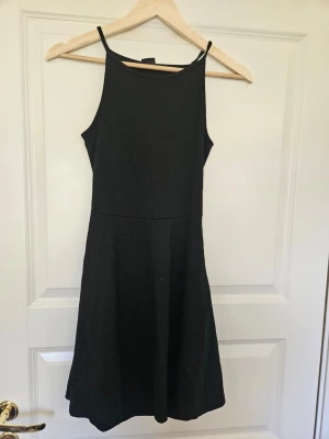 Klänning Gina Tricot S - En enkel och stilren svart klänning med smala axelband och rundad halsringning. Klänningen har en lätt utställd kjol och är ärmlös, vilket ger en clean och modern look. Perfekt för dig som gillar minimalistisk stil.  E4