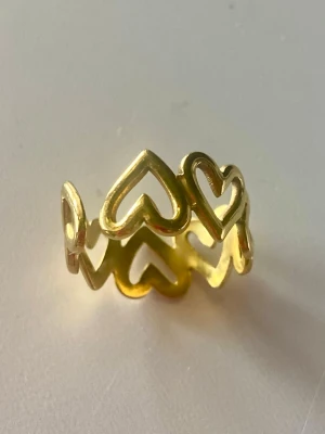 Guldfärgad ring med hjärtan - Säljer en snygg guldfärgad ring med öppet mönster av hjärtan runt hela ringen. Ringen har en modern och lekfull design och är justerbar i storlek. Perfekt för dig som gillar unika accessoarer med romantisk touch.