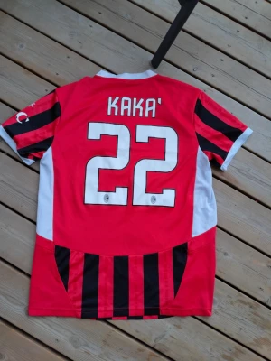 AC Milan matchtröja Kaka #22 - Snygg AC Milan matchtröja med Kaka och nummer 22 på ryggen. Tröjan är röd och svart med vita detaljer, klassiska vertikala ränder och klubbmärke på bröstet. Emirates Fly Better som sponsortryck på framsidan. Perfekt för fotbollsfans!