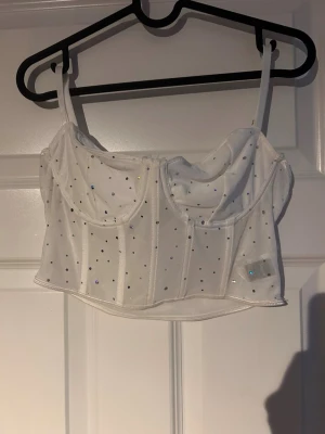 Vit glittrig korsettopp  - Säljer en vit korsettopp med tunna axelband och glittriga strassdetaljer.Perfekt att styla med höga jeans eller kjol 