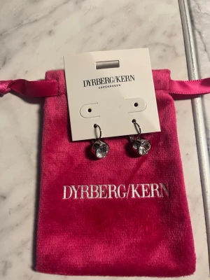 Silverörhängen med kristall från Dyrberg/Kern - Snygga örhängen från Dyrberg/Kern i silverfärg med rund, klar kristall som hänger i en enkel krok. Kommer i en lyxig rosa sammetsförpackning med logga. Perfekta för dig som gillar stilrena och glittriga accessoarer.