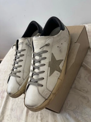 Golden Goose Super stars  - Snygga vita sneakers från Golden Goose med grå stjärna på sidan och grå skosnören. Skorna har en låg silhuett och detaljer i svart vid hälen. Tillverkade i läder med slitna detaljer för en cool, avslappnad look.