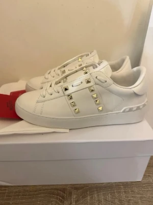Vita Valentino sneakers med nitar - Snygga vita sneakers från Valentino Garavani med coola guldfärgade nitar på sidorna och över vristen. Skorna är i läder och har en klassisk låg siluett med vita snören och diskret mönster på hälen. Perfekta för dig som vill sticka ut med stil.
