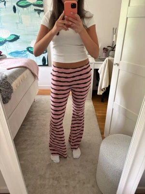 Rosa stickade loungewear-byxor - Säljer ett par mysiga stickade loungewear-byxor i rosa med svarta vågiga ränder. Byxorna har utsvängda ben och hög midja, perfekta för chill hemma. Materialet ser ut att vara mjukt och stretchigt, vilket gör dem superbekväma att ha på sig.