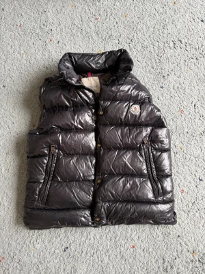 Svart dunväst från Moncler - Snygg svart dunväst från Moncler med glansig finish och klassisk logga på bröstet. Västen har hög krage, två dragkedjeförsedda fickor och tryckknappar framtill. Perfekt för lager-på-lager och streetstyle. Materialet är dunfyllning och yttertyget är glansigt syntet.
