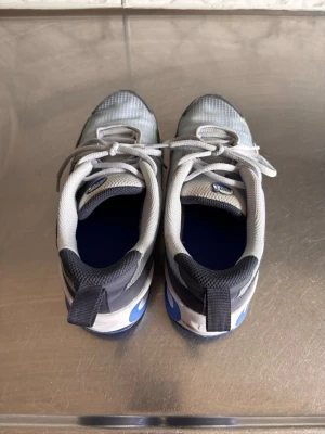 Nike grå/blå sneakers med mesh - Nike sneakers i grått och blått med vita detaljer. Ovandelen är i mesh för bra ventilation och har klassisk snörning. Skorna har lågt skaft och en sportig look med blå detaljer på hälen och vit sula. Perfekta för dig som gillar streetwear och sportig stil.