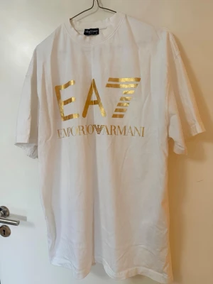 Vit t-shirt - Snygg vit t-shirt med stort guldfärgat tryck framtill och mindre. Klassisk passform och rund hals. Tillverkad i mjuk bomull som känns skön mot huden. Perfekt för dig som gillar stilrena och exklusiva plagg.