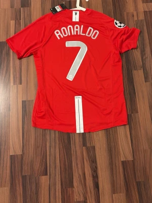Manchester United 2007-8 Ronaldo Retro Tröja - Säljer en röd Manchester United matchtröja från Nike med Ronaldo och nummer 7 på ryggen. Tröjan har Champions League-märke på ärmen, AIG som sponsor på bröstet och klubbmärke fram. Tillverkad i lätt och ventilerande material, perfekt för fotboll eller samling.
