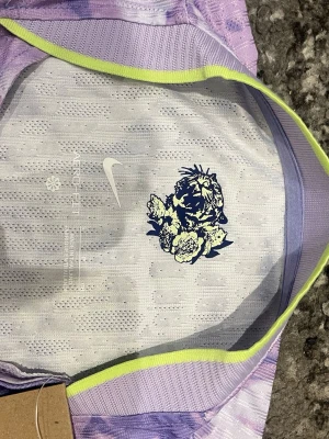 Lila blommig fotbollströja Nike Korea - Säljer en snygg lila och vit fotbollströja från Nike med blommigt mönster och Syd Korea-logga på bröstet. Tröjan har korta ärmar, rund hals och är tillverkad i ett lätt och ventilerande polyestermaterial. Perfekt för dig som gillar unika sportkläder!