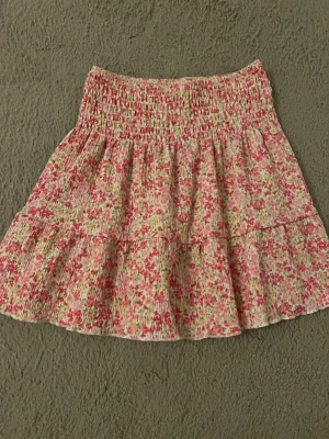Blommig volangkjol i rosa toner Gina tricot  - Söt kort kjol med volanger och smock i midjan. Tyg i ljusrosa, vitt och grönt med småblommigt mönster. Kjolen har en luftig och flowy känsla, perfekt för varma dagar. Materialet känns lätt och mjukt.