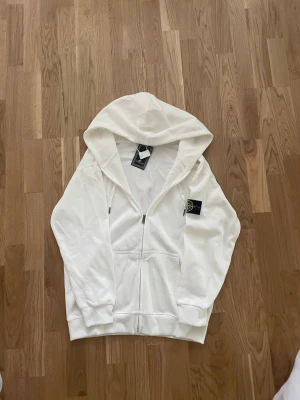 Vit hoodie från Stone Island - Säljer en clean vit hoodie från Stone Island med dragkedja framtill och klassisk logopatch på ärmen.  Hoodien  är i XL men är väldigt liten strolek på tröjan och kan därför passa storlekarna L och M.
