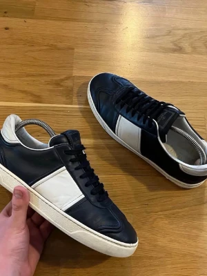 Svarta och vita sneakers från Valentino - Snygga svarta sneakers från Valentino med vita detaljer och vit sula. Skorna har en låg profil, svarta skosnören och coola nitar på hälen. Tillverkade i läder för en lyxig känsla och stilren look.