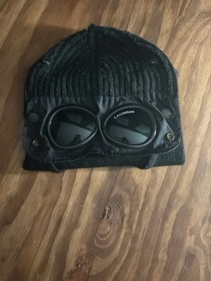 Svart balaclava med glasögon Goggle - Säljer en svart stickad balaclava från C.P. Company med inbyggda goggle-glasögon och detaljer i nylon. Balaclavan har öppning för ansiktet och coola metallknappar vid sidorna. Perfekt för dig som vill sticka ut med streetwear-vibe.