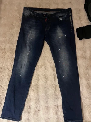 Mörkblå skinny jeans med slitningar - Säljer ett par mörkblå skinny jeans med slitningar och färgstänk för en trendig, avslappnad look. Jeansen har fem fickor, knappgylf och tydliga tvättade partier framtill. Perfekta för dig som gillar streetstyle och vill ha något unikt i garderoben.
