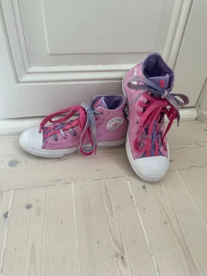 Rosa Converse high top sneakers - Säljer ett par rosa Converse high tops med lila och rosa detaljer. Skorna har dubbla skosnören i rosa och lila, vit tåhätta och klassisk Converse-logga på sidan. Insidan är lila och materialet är canvas. Perfekta för dig som vill sticka ut med färg!